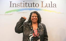 Ivone Silva, uma mulher negra, de cabelos escuros e cacheados, posa com o punho erguido em frente a um painel com o logo do Instituto Lula